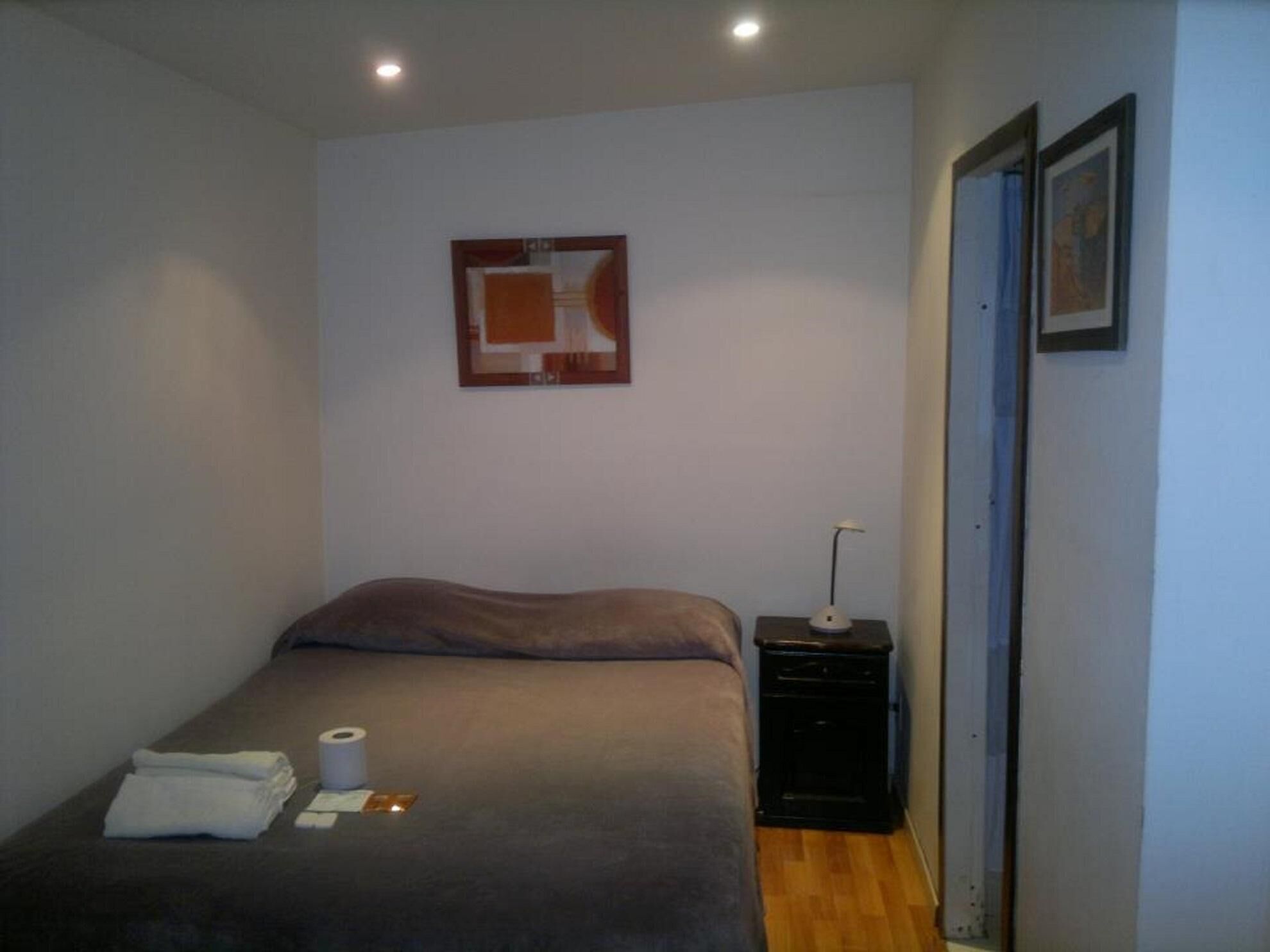 A small, sparsely furnished bedroom with a bed, a nightstand, a lamp, and a framed picture on the wall.