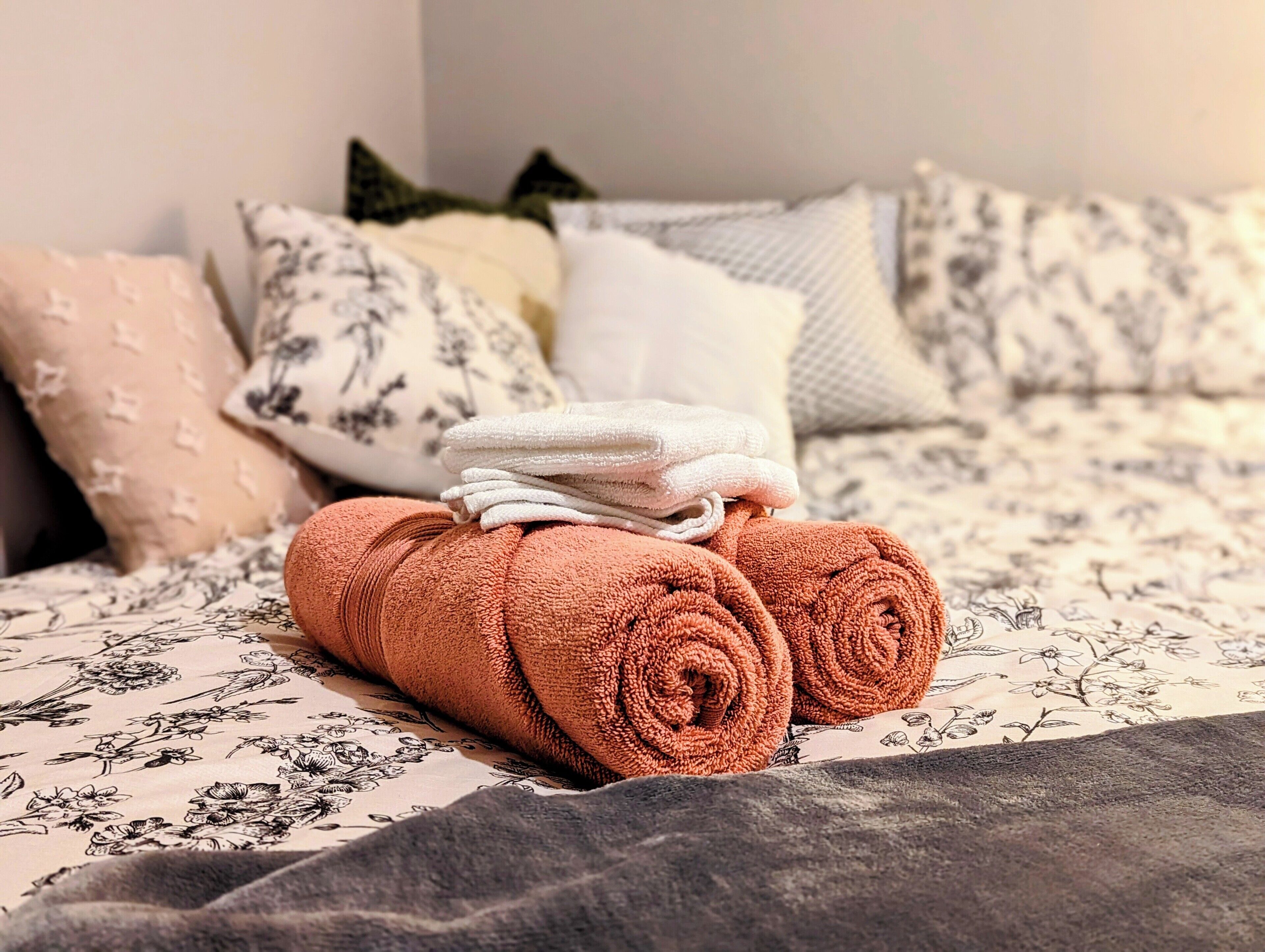 A neatly made bed with a floral-patterned bedspread, a stack of rolled towels, and several decorative pillows.