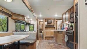 Interior de una caravana con zona de comedor, kitchenette y ventana con vistas a árboles.