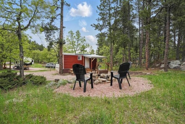 Une petite cabane aux murs rouges, entourée d’une forêt. On peut voir deux chaises noires devant la cabane.