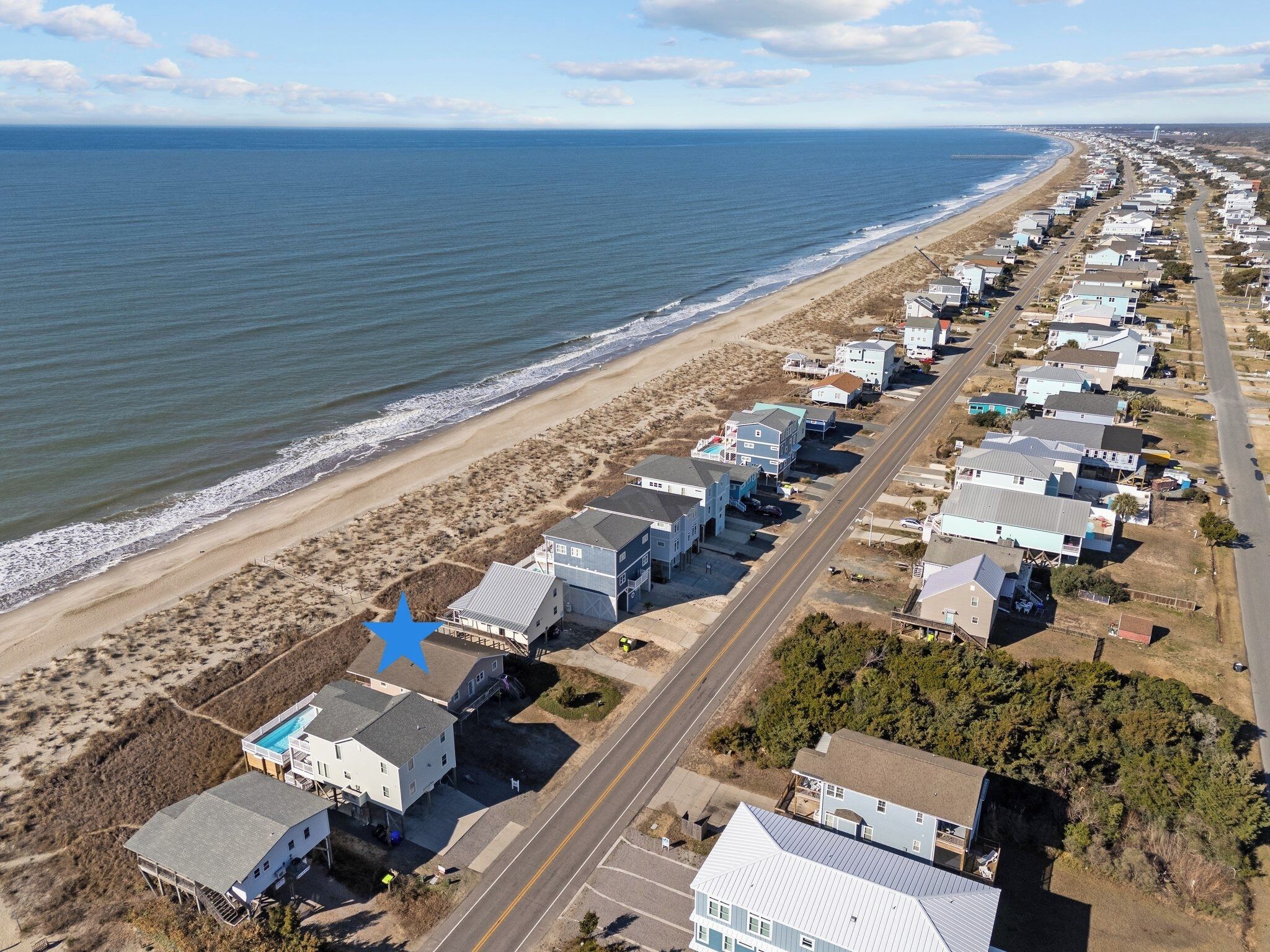 A coastal residential area with houses along the beach, a road, and a clear view of the ocean.