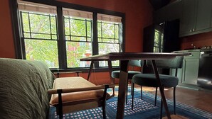 Een kamer met een bed, een tafel en een stoel. Er zijn ramen met uitzicht op bomen en gebouwen. Een keuken is op de achtergrond zichtbaar.