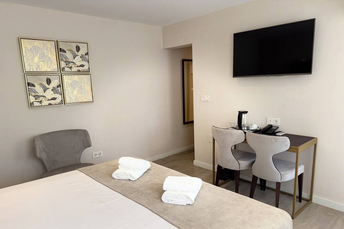 A hotel room with a bed, a desk with two chairs, a flat-screen TV, and artwork on the wall.