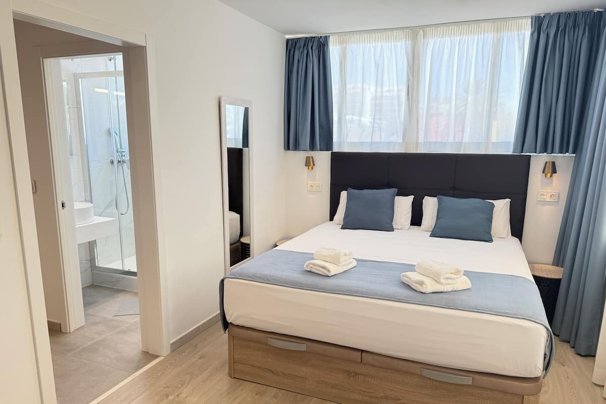A modern hotel room with a bed, bedside tables, a mirror, and a view of the outside through the window.