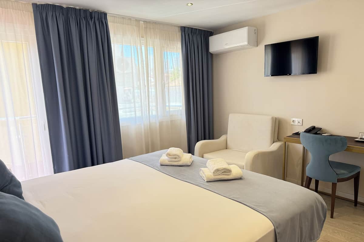 A hotel room with a large bed, a desk with a chair, a TV mounted on the wall, and an air conditioning unit.