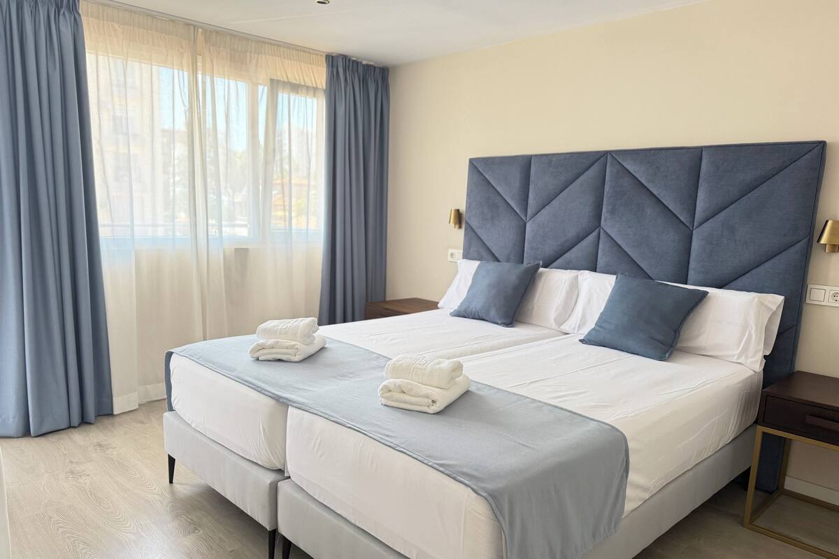A double bed with a tufted headboard, two pillows, and a folded towel on each side.