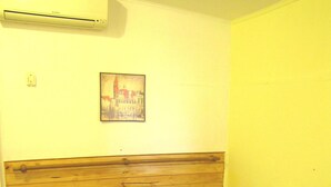 Een kleine, eenvoudige kamer met een houten hoofdbord, een enkel bed met een geruite deken, een nachtkastje met een lamp en een wandairconditioner.