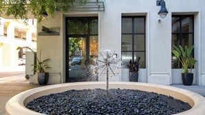 Une fontaine circulaire avec une sculpture centrale, entourée d’une bordure en pierre, située devant un bâtiment doté de grandes fenêtres et d’une porte.