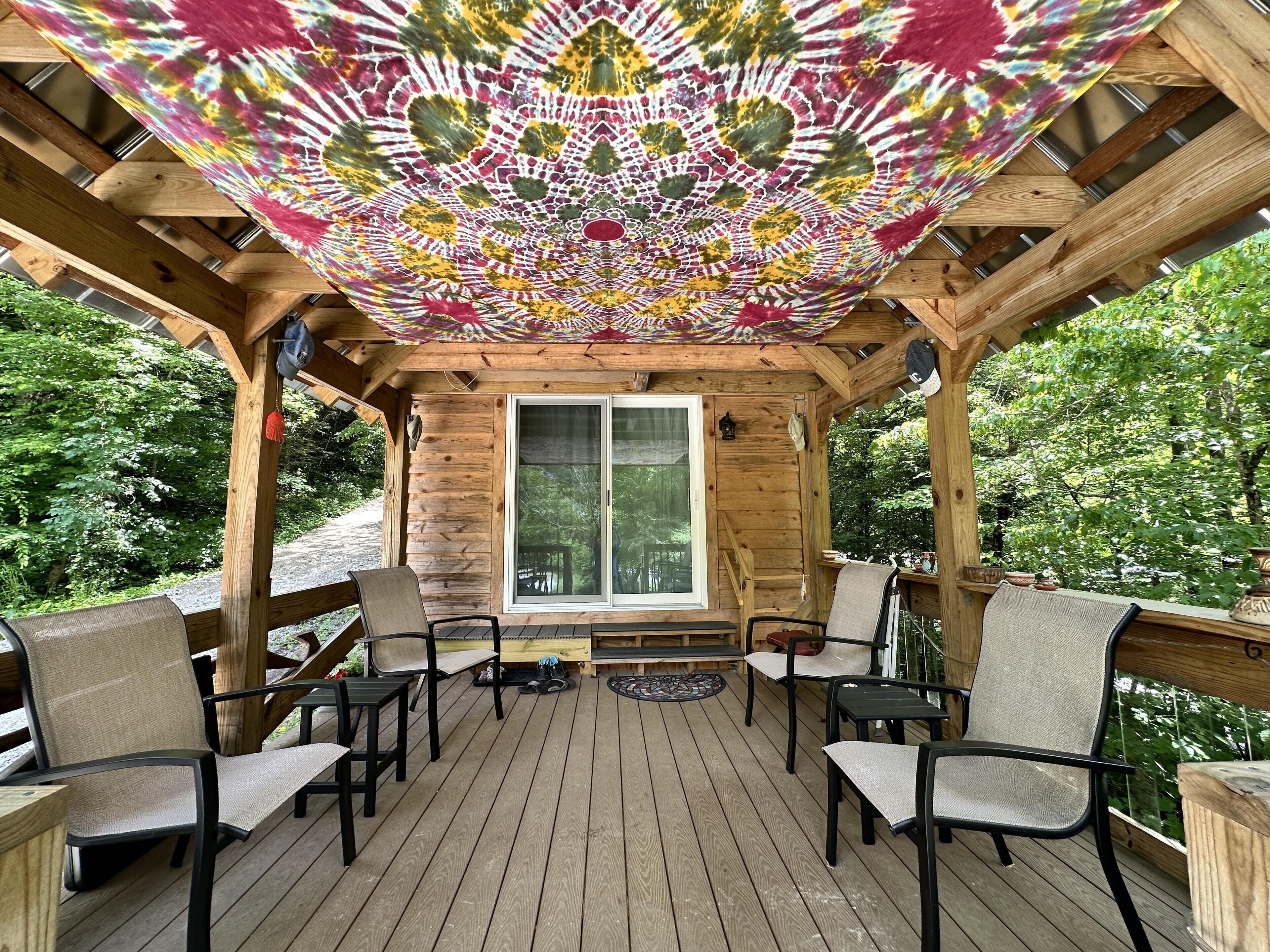 A wooden deck with a covered seating area, a large window, and a view of the surrounding greenery.