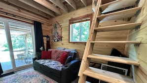 A cozy cabin interior with a wooden staircase, a sofa with a patterned throw, and a large window overlooking greenery.