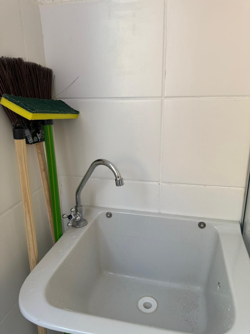 Un lavamanos blanco con grifo cromado, una escoba verde y un cepillo con mango amarillo frente a una pared de azulejos blancos.