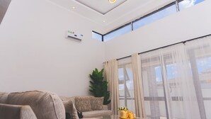 A modern living room with a sofa, a coffee table with decorative items, a potted plant, and a large window with curtains.