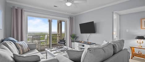 A modern living room with a grey sofa, a coffee table, a flat-screen TV, and a balcony with a view.