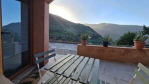 Ein Balkon mit einem Holztisch und Stühlen, Blumentöpfen und Blick auf grüne Hügel im Hintergrund.
