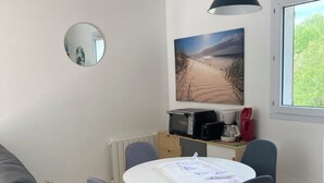 A small dining area with a round table, four chairs, a black pendant light, a kitchen area with appliances, a large window, and a beach scene painting on the wall. - Bright vacation home in the dunes, kerhillio beach (Erdeven)