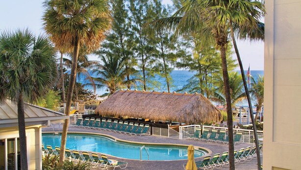 A pool area with a thatched-roof structure, surrounded by palm trees and a view of the ocean.