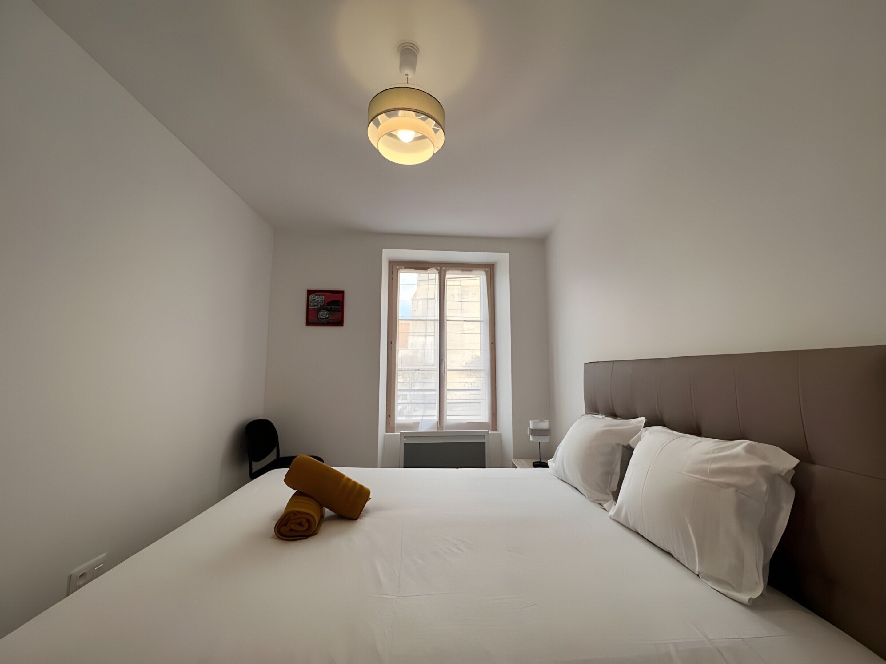 Un dormitorio con una cama grande, una ventana, una lámpara en el techo y un pequeño letrero rojo en la pared.