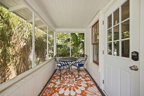 A covered porch with a table and chairs, a door with a window, and a view of greenery.