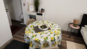 A compact living space with a round dining table covered in a lemon pattern, a small side table with a lamp, a desk with a potted plant, and a bed with a patterned bedspread. - Studio Apartment Enter - Studio Apartment With Terrace (Karlovac)