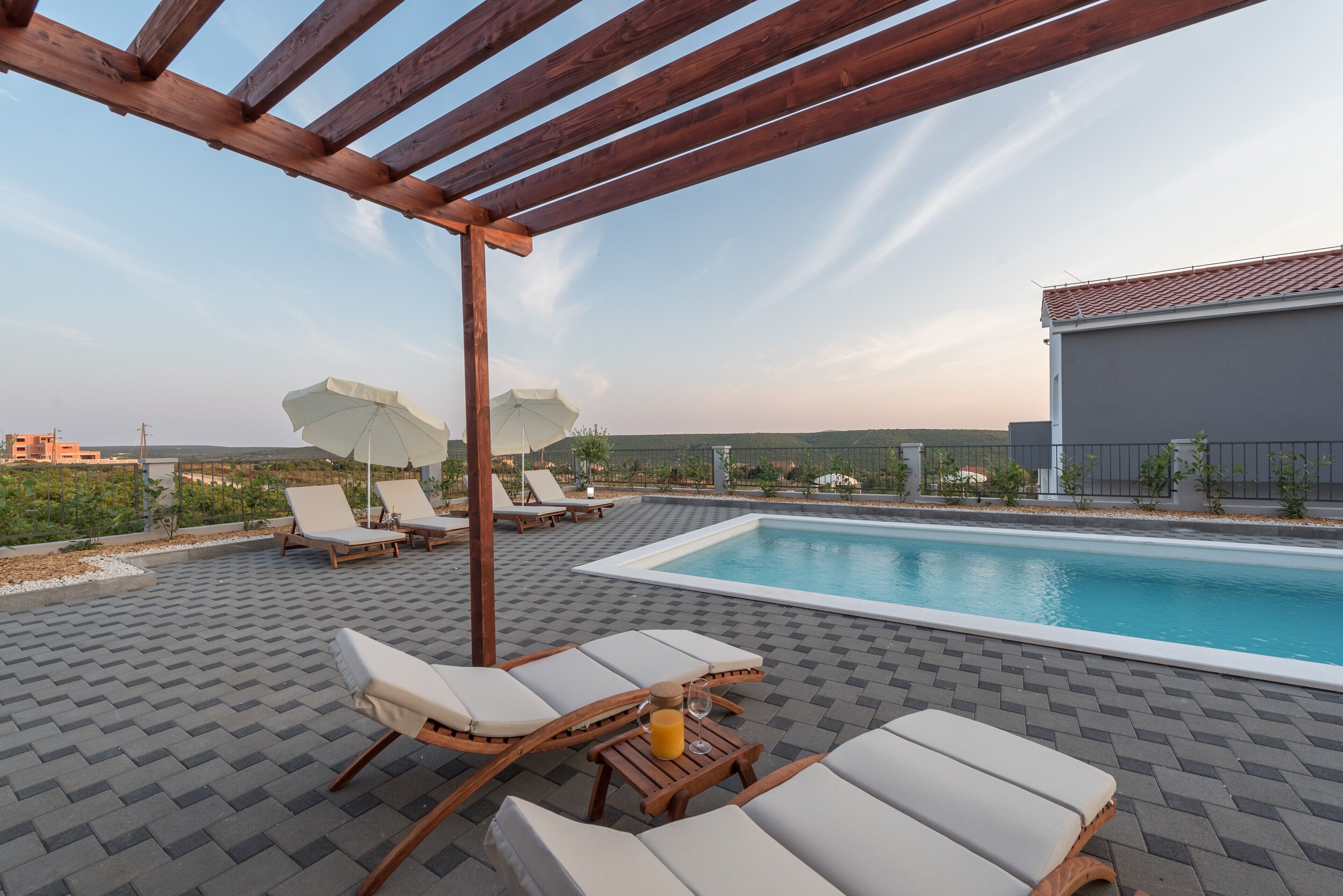 A rooftop pool area with lounge chairs and umbrellas, a wooden pergola, and a view of the cityscape.