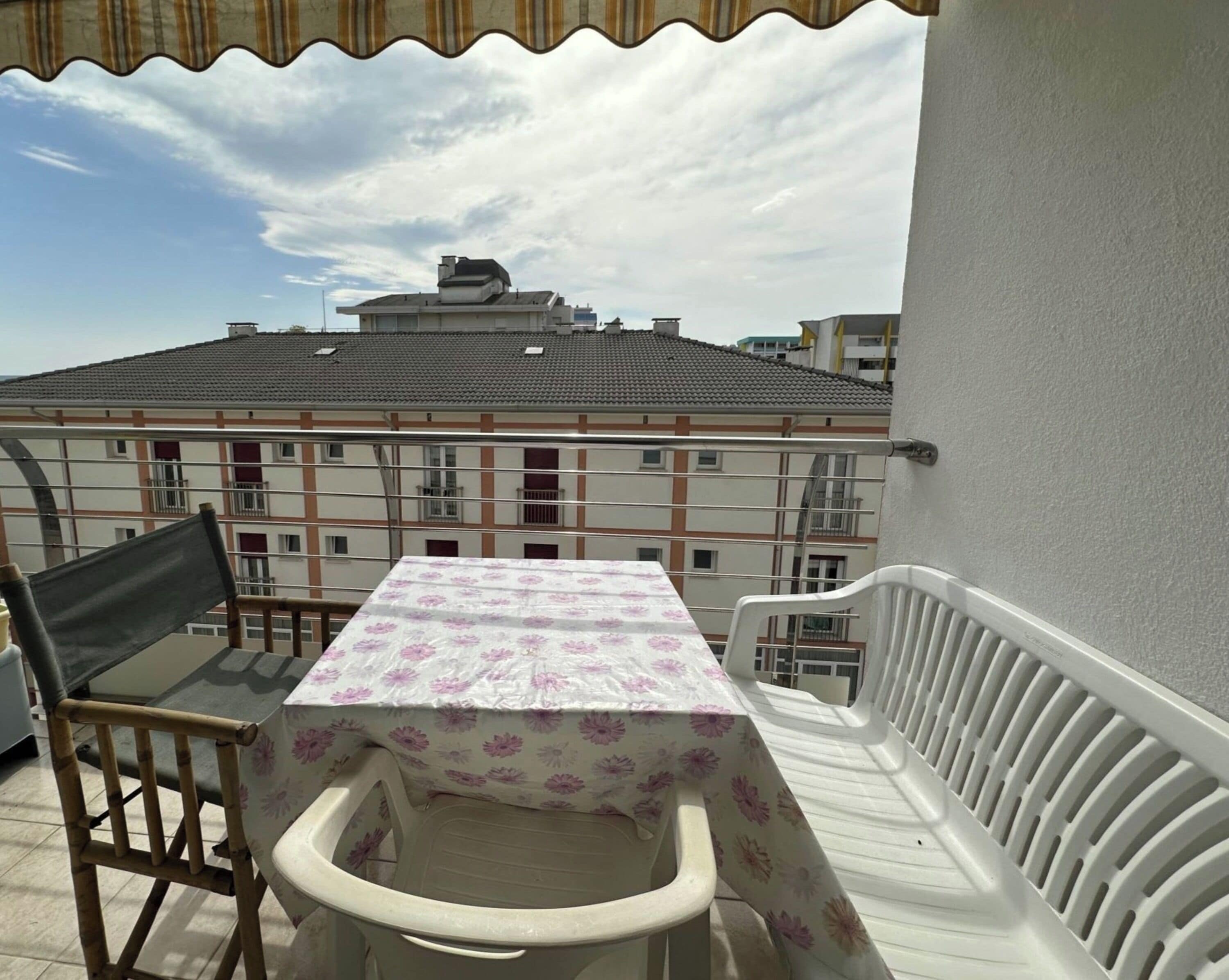 Uma varanda com mesa e cadeiras, um corrimão branco e vista para edifícios ao fundo.