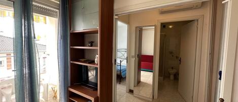 A spacious room with a large wooden bookshelf, a sliding glass door leading to a balcony, and a view of a red bed in the adjacent room.