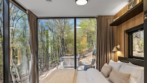 Un dormitorio moderno con una cama grande, vistas a árboles a través de ventanales de piso a techo, y un estante empotrado con una lámpara.