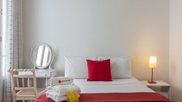 Ein ordentlich eingerichtetes Hotelzimmer mit einem Bett, einem Nachttisch mit Lampe, einem Sessel, einem Spiegel und einem kleinen Tisch mit Handtüchern und Toilettenartikeln.
