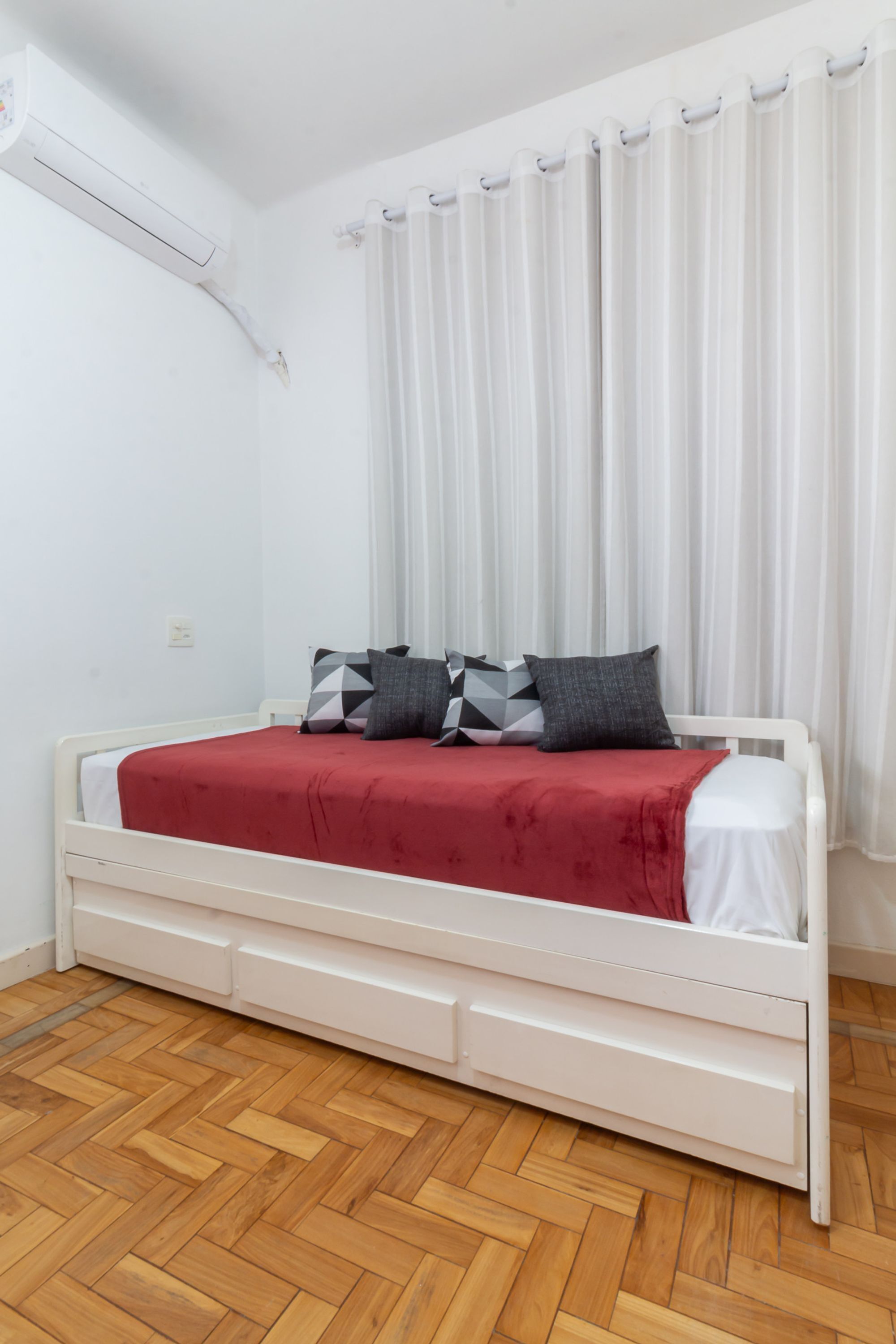 A white bed with a red blanket, wooden floor, white walls, and a white air conditioning unit.