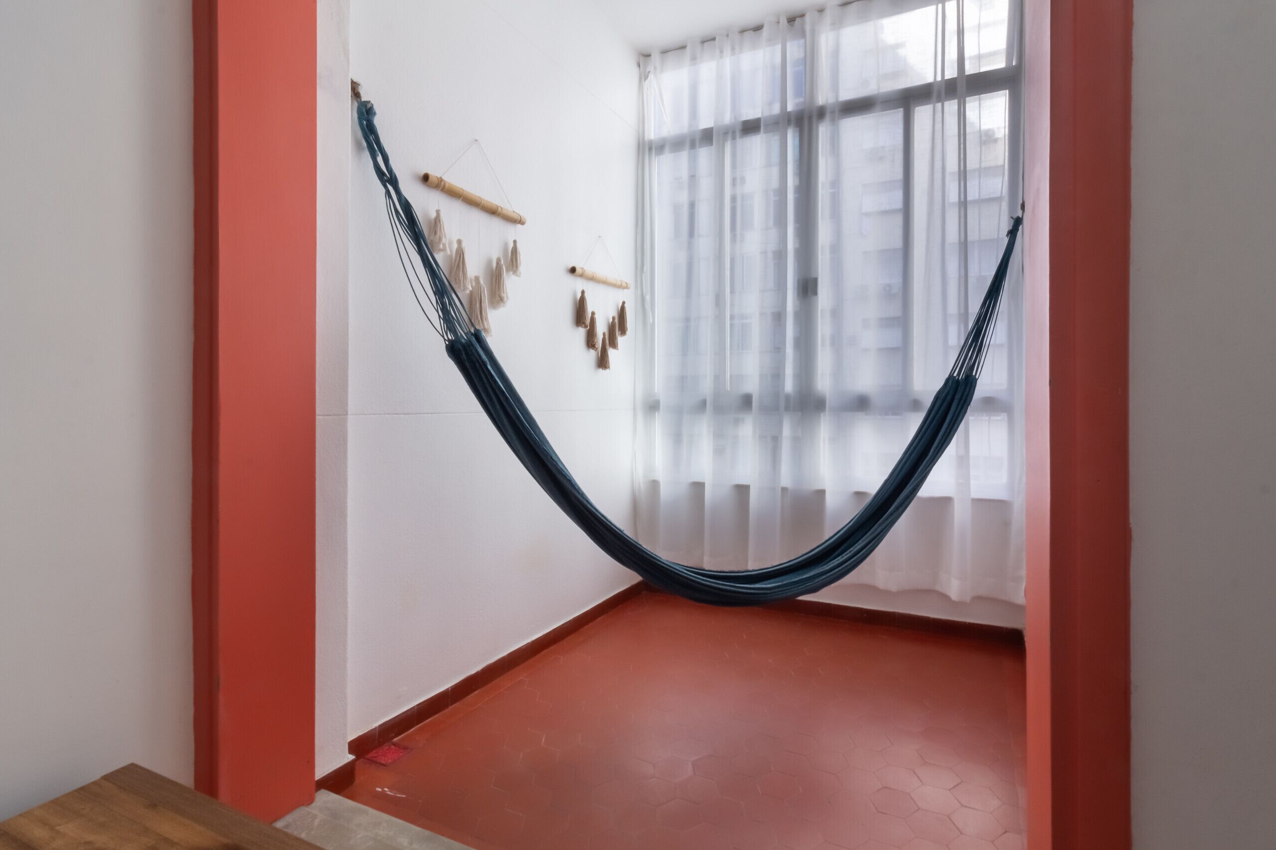 Una hamaca colgada en una habitación con piso rojo, paredes blancas y una ventana con cortinas traslúcidas.