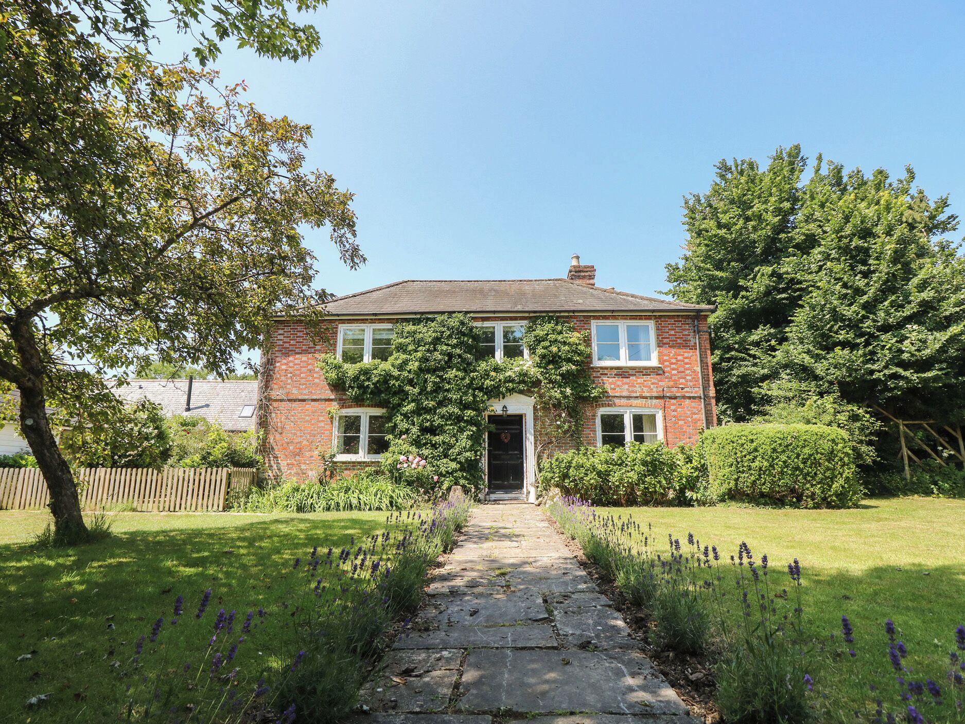 A brick house with a stone pathway leading to the front door, surrounded by greenery and lavender plants.
