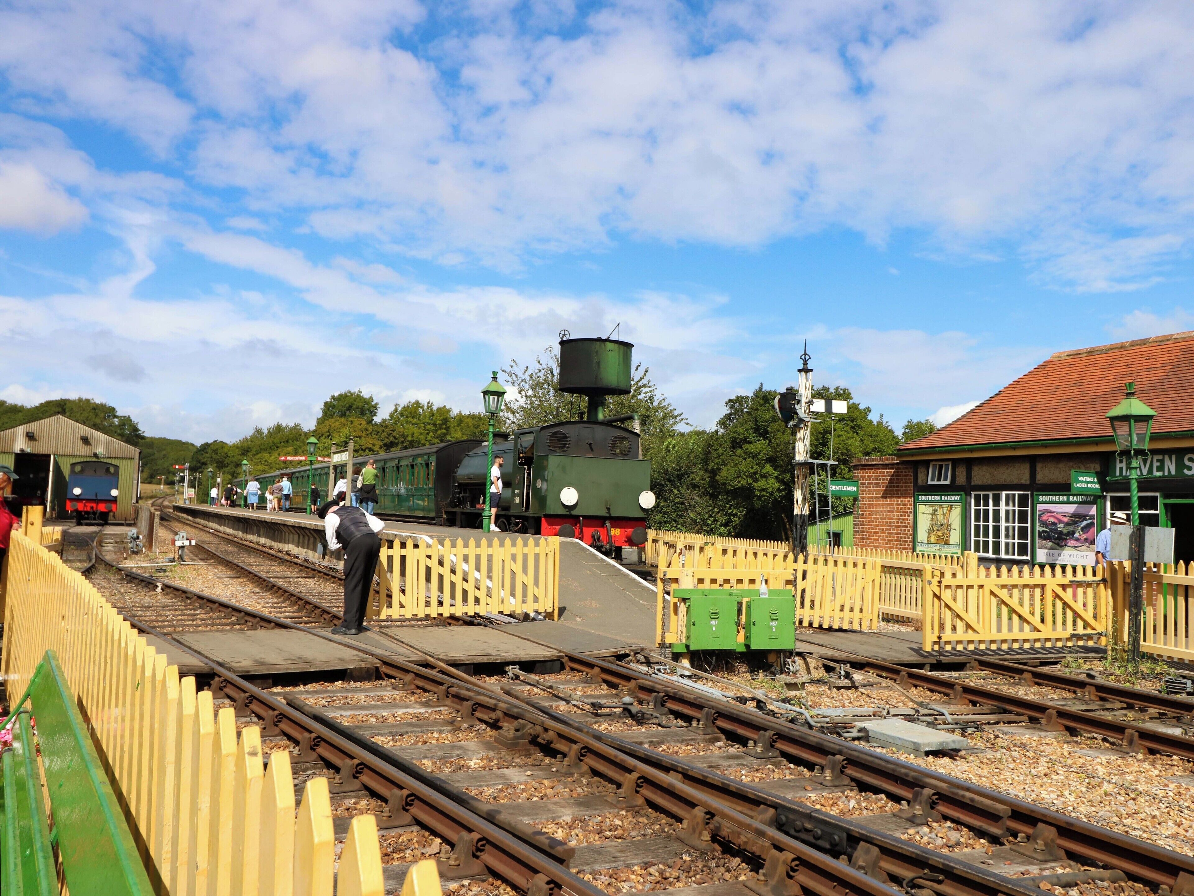 Un andén de estación de tren con varias vías, un tren verde y un edificio con un letrero que dice 'Haven'.