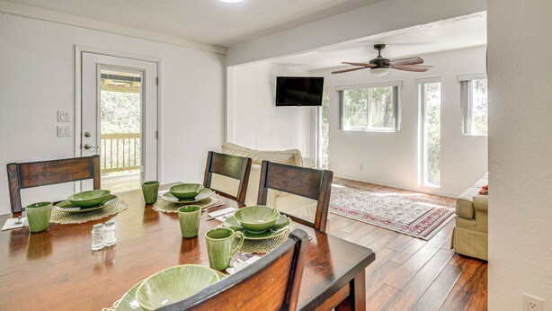 A dining area with a wooden table set for four, surrounded by chairs. A television is mounted on the wall, and there are windows providing natural light. - Walk to Lake George: Quiet Nature Escape w/ Deck (Fort McCoy)