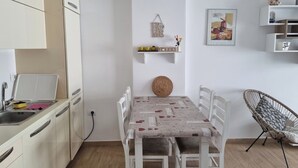 Una cocina compacta con armarios blancos, una mesa de comedor con sillas, una lámpara colgante y una estantería con objetos decorativos.