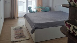 Un dormitorio con cama, una mesita, sillas, una planta colgante y una ventana con cortinas.