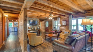 A log cabin interior with a wooden ceiling, a stone fireplace, a television, and a cozy seating area with leather sofas and armchairs. - Waterfront Cabin on Lake Sinclair w/ Private Dock! (Eatonton)