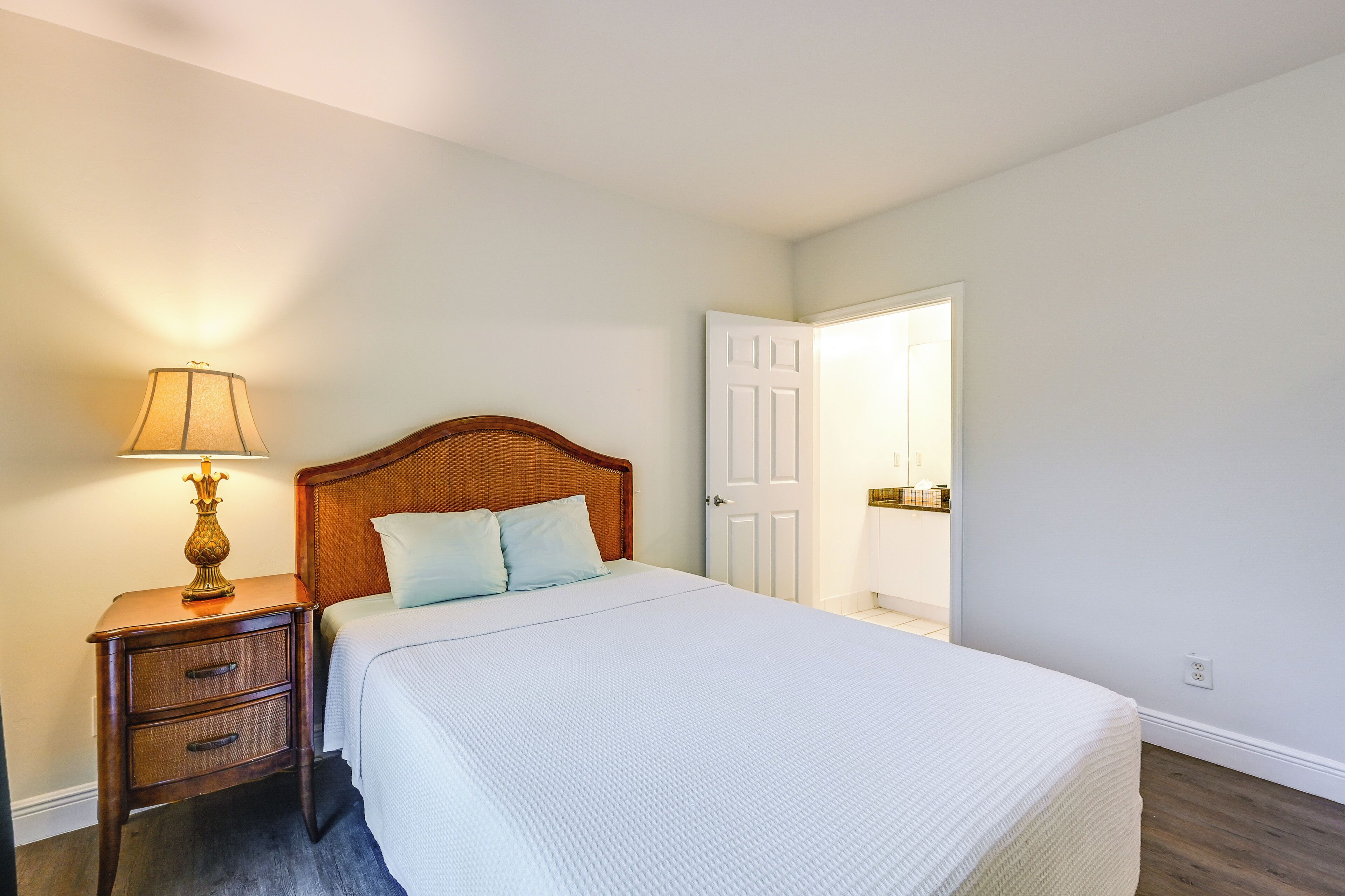 Une chambre à coucher avec une tête de lit en bois, une lampe de chevet, un couvre-lit blanc et une porte donnant sur une autre pièce.