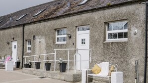 Une rangée de bâtiments en pierre dotés de nombreuses fenêtres et portes, un banc blanc avec un coussin jaune et des plantes en pot le long du trottoir.