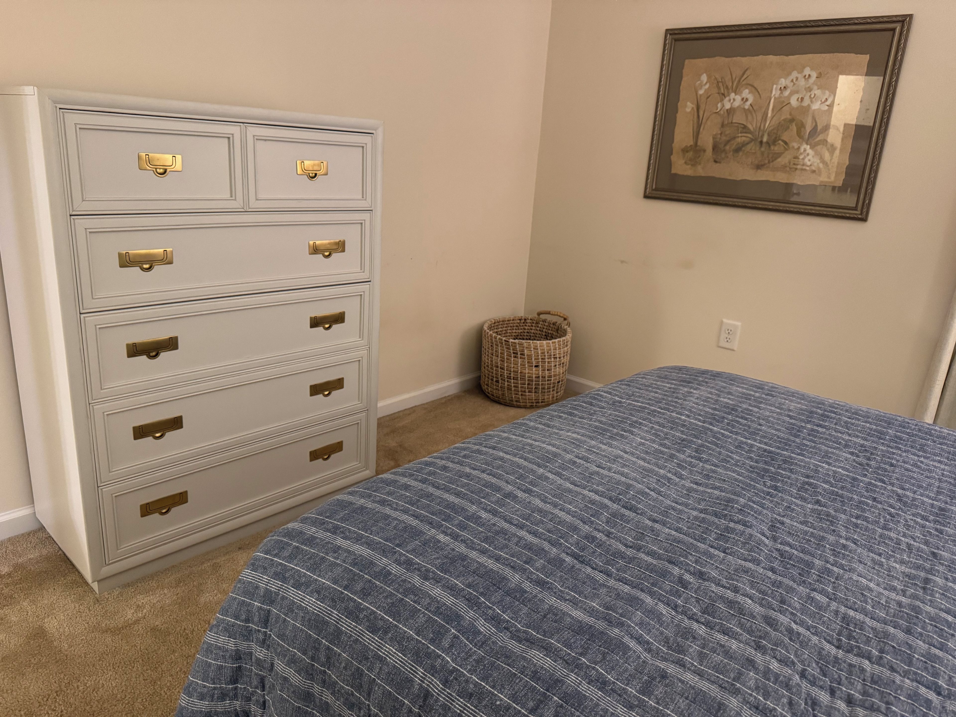 Una camera da letto con un letto, una cassettiera, un cesto in vimini e un quadro appeso al muro.