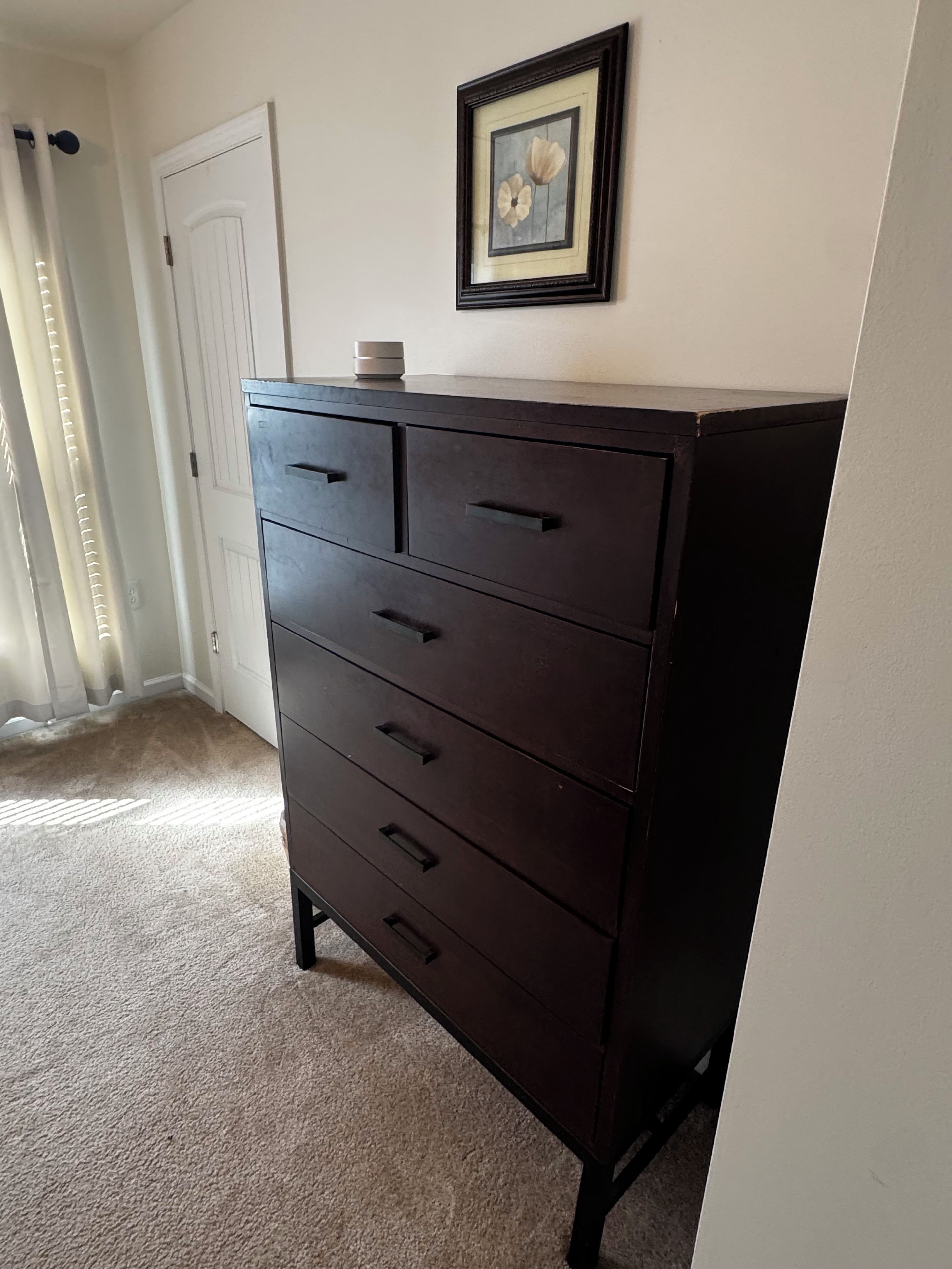 A dark wooden dresser with multiple drawers, a framed picture on the wall, and a white door in the background.