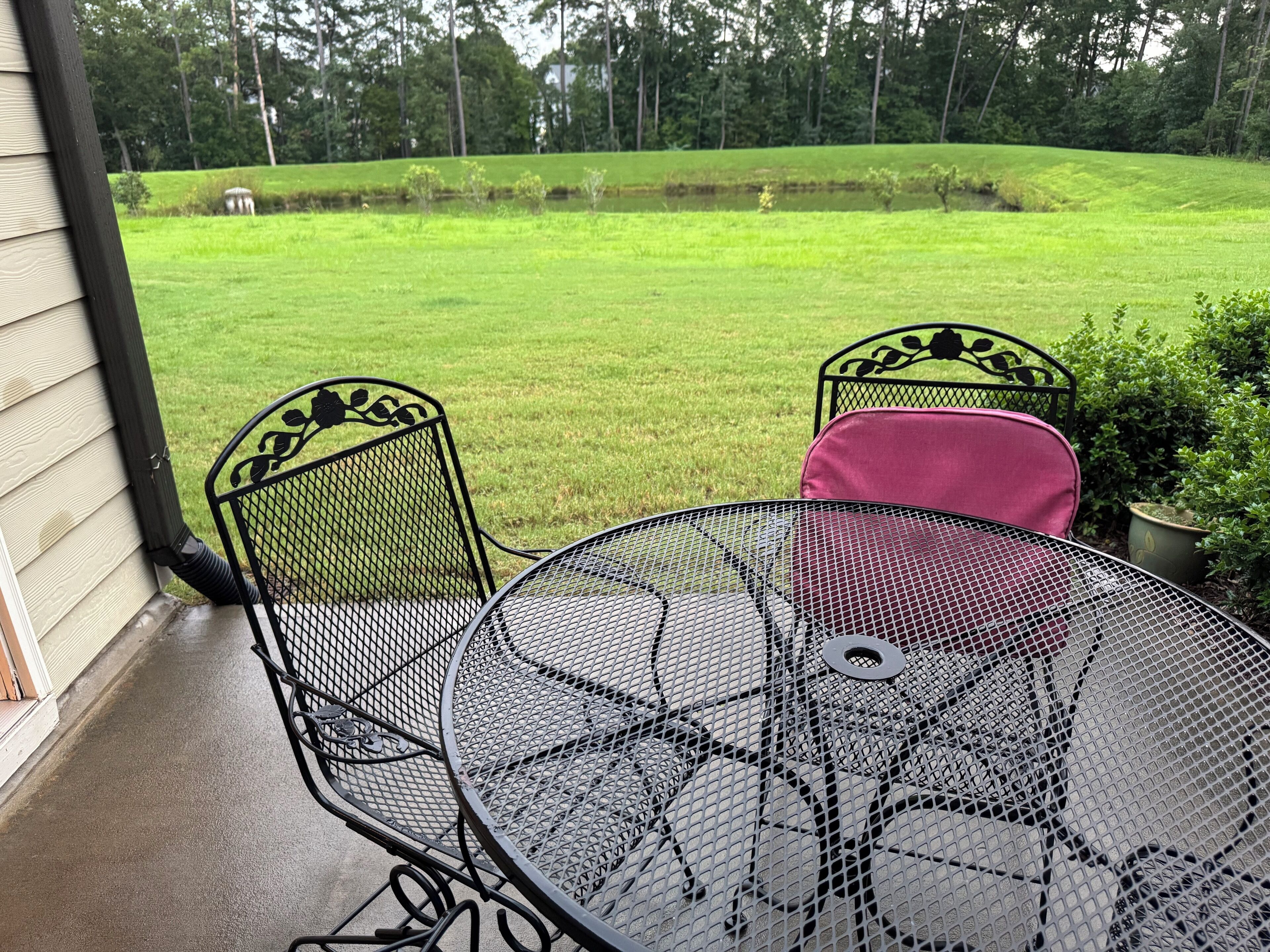 Un patio con una mesa redonda de metal y dos sillas, con vista a un jardín con árboles.