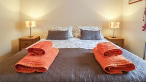Uma cama bem arrumada com duas almofadas, duas mesas de cabeceira com abajures e duas toalhas enroladas sobre a cama.