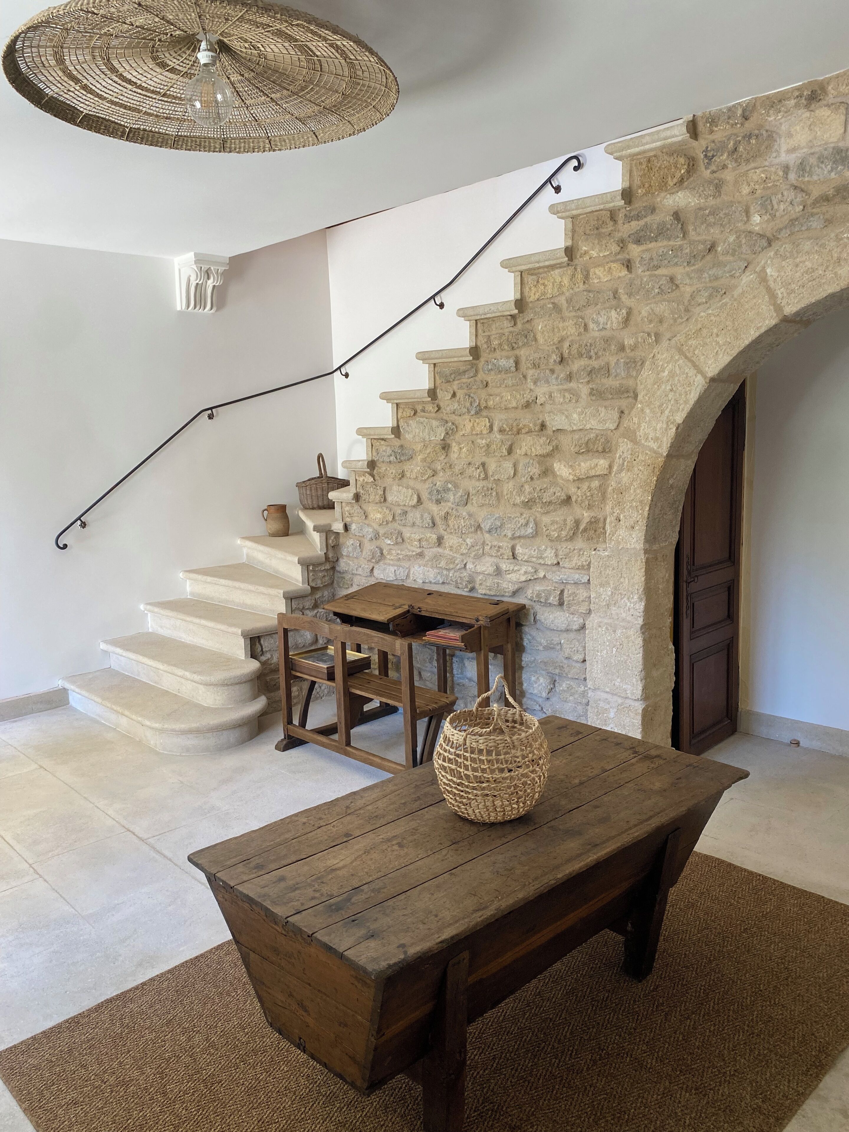 Un escalier en pierre menant à une porte en bois, un luminaire tressé suspendu au-dessus, et une table en bois rustique sur laquelle repose un panier tressé.