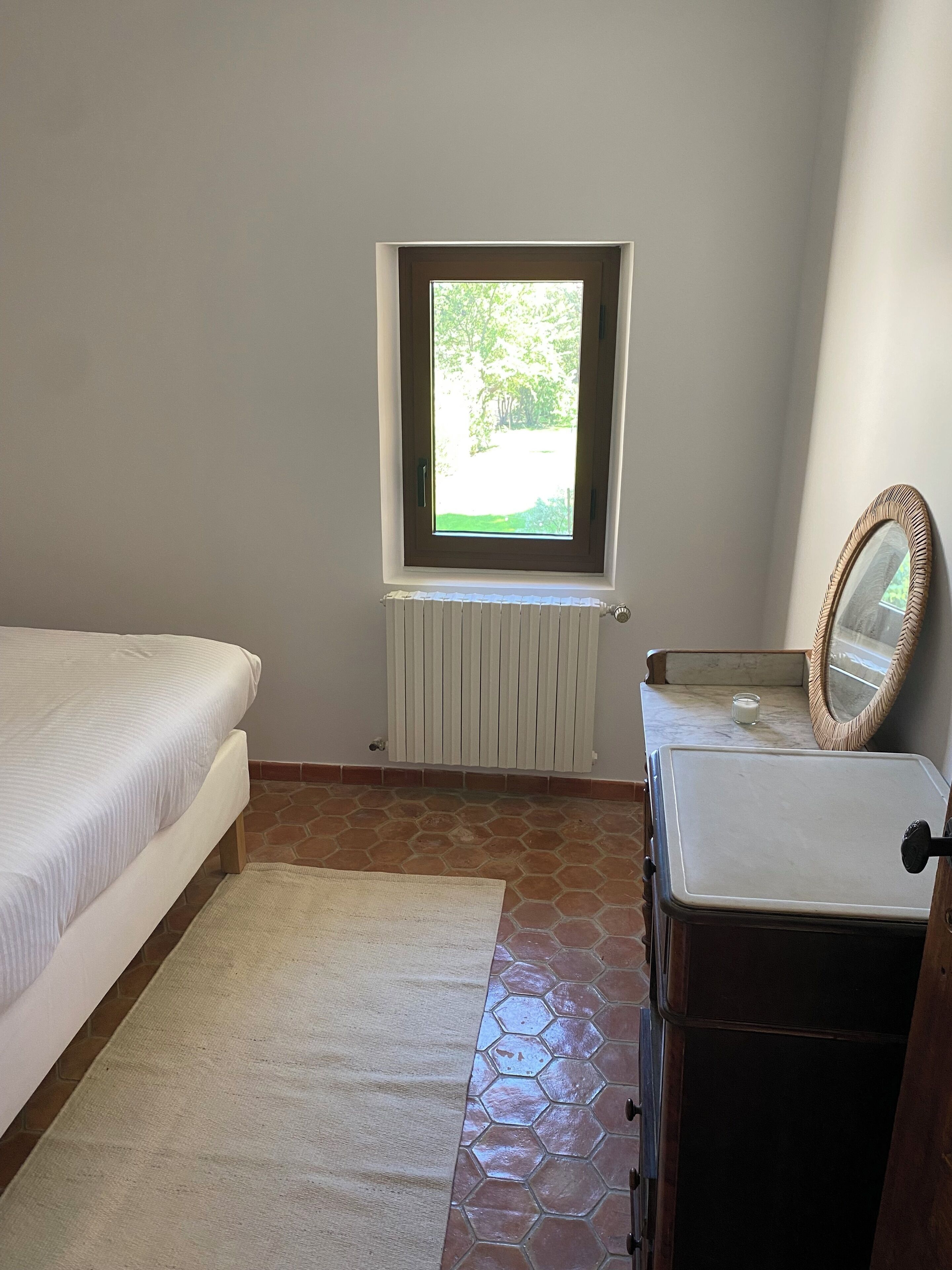 Ein Schlafzimmer mit Bett, Fenster, Heizkörper, Spiegel und Kommode.