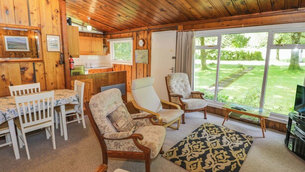 L’intérieur chaleureux d’un chalet avec des murs en bois, un coin repas, deux fauteuils, un canapé et une grande fenêtre donnant sur une cour verdoyante.