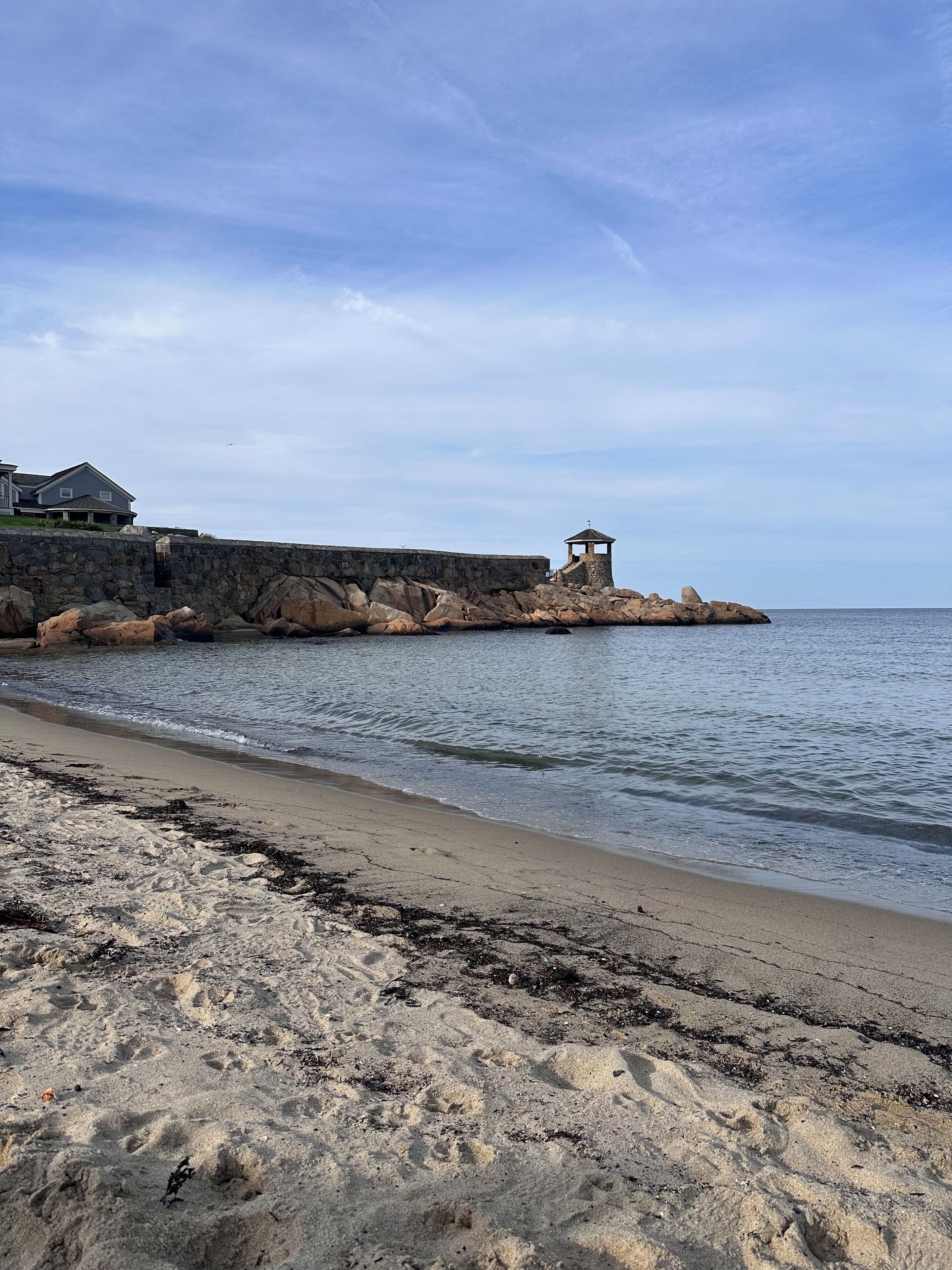 A coastal scene with a stone pier leading to a small gazebo, a sandy beach, and a house in the background.