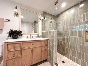 A bathroom with a glass-enclosed shower, a wooden vanity with drawers, a large mirror, and a potted plant on the vanity.