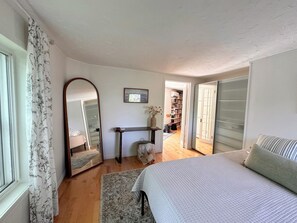 Un dormitorio con cama, espejo, mesita de noche, ventana con cortinas, puerta y un pasillo que conduce a una biblioteca.