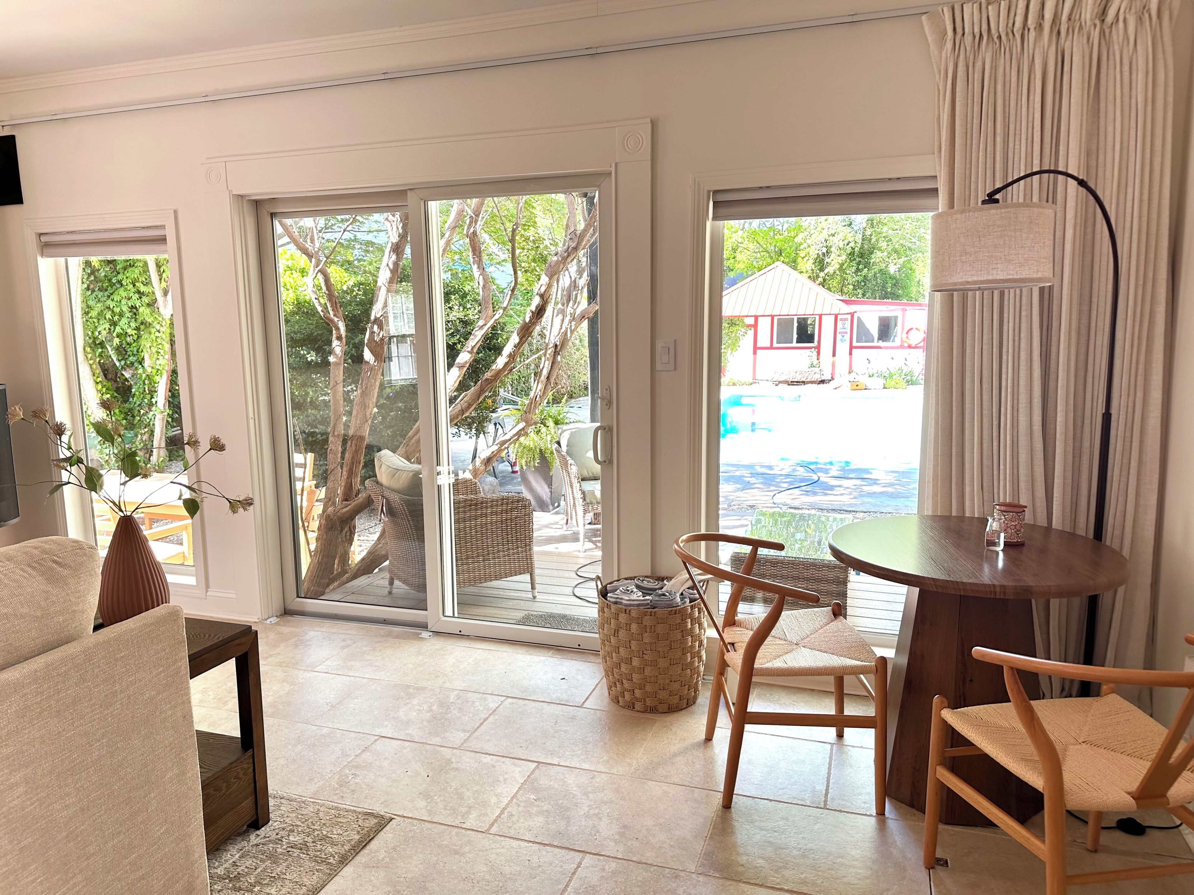 Une pièce comprenant un canapé, une table à manger avec des chaises, un panier en osier et une vue, par une porte-fenêtre coulissante, sur un espace extérieur avec un bâtiment rouge.