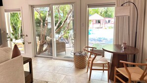 A room with a sofa, a dining table with chairs, a wicker basket, and a view through a sliding glass door to an outdoor area with a red building. - Newly Updated! Private Pool! Walk to Beach, Shops, Restaurants & Train to Salem! (Rockport)